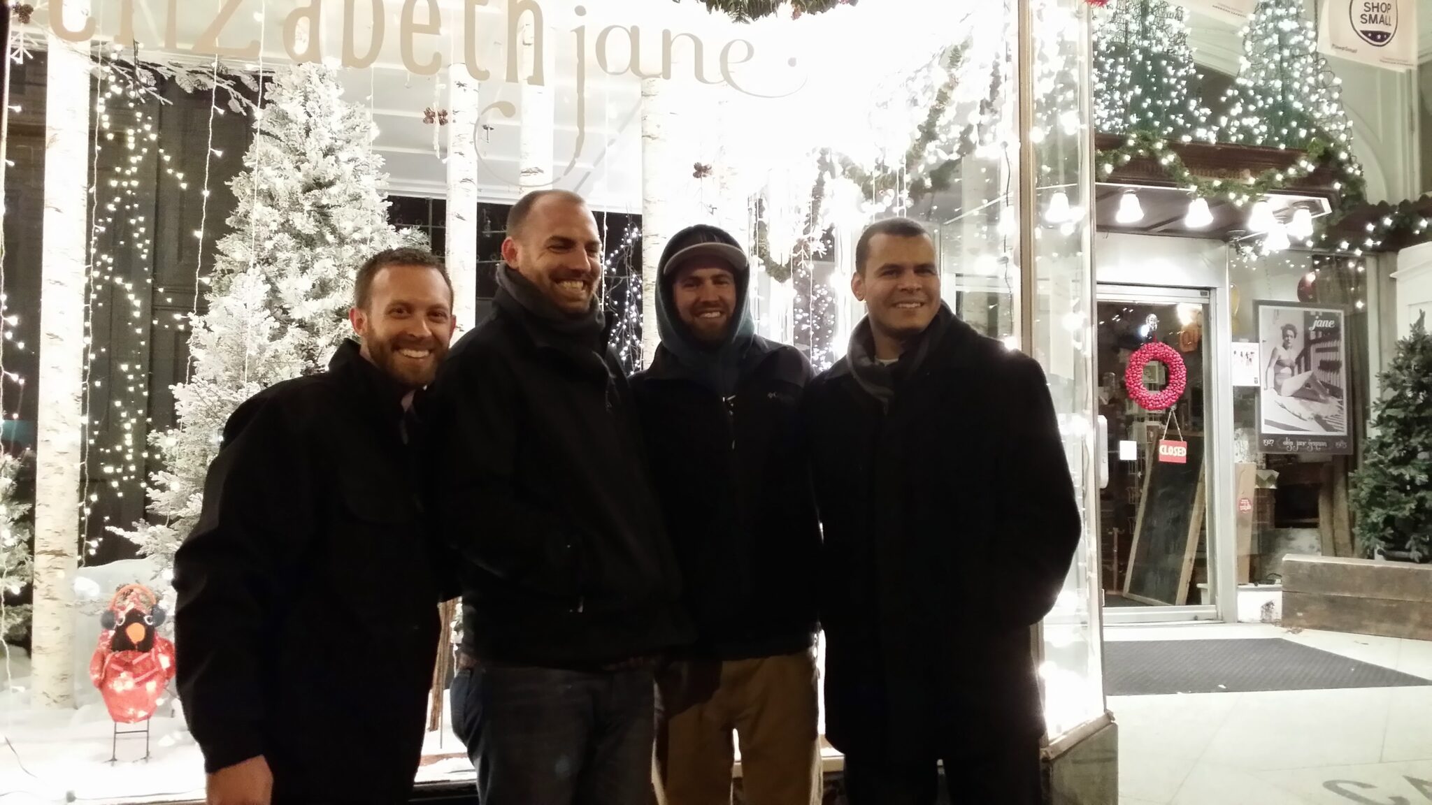 Four men wearing winter coats smile and stand close together for a group picture outside in front of a storefront decorated for the holidays with lights