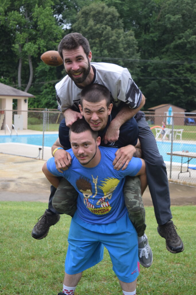 Three men, each one piggybacked on top of the other, pose smiling for a picture in front of a pool