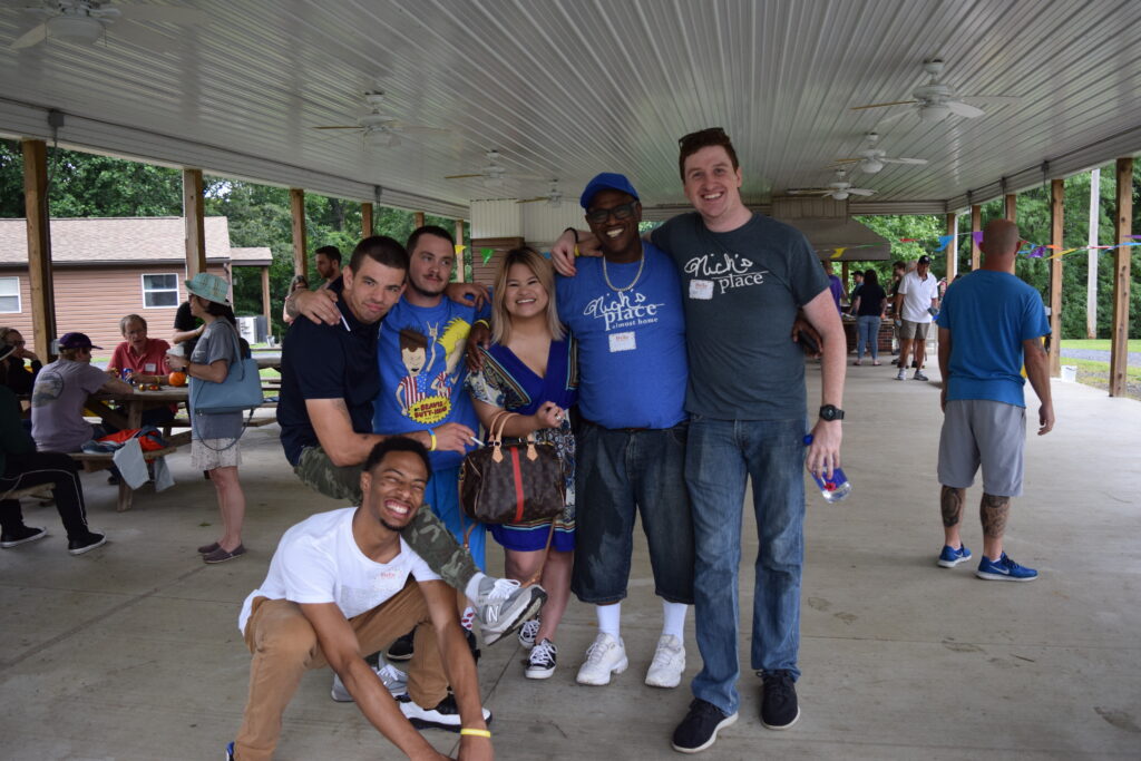 A group of five men and one woman stand together smiling and posing for a picture at an outdoor party under a pavilion