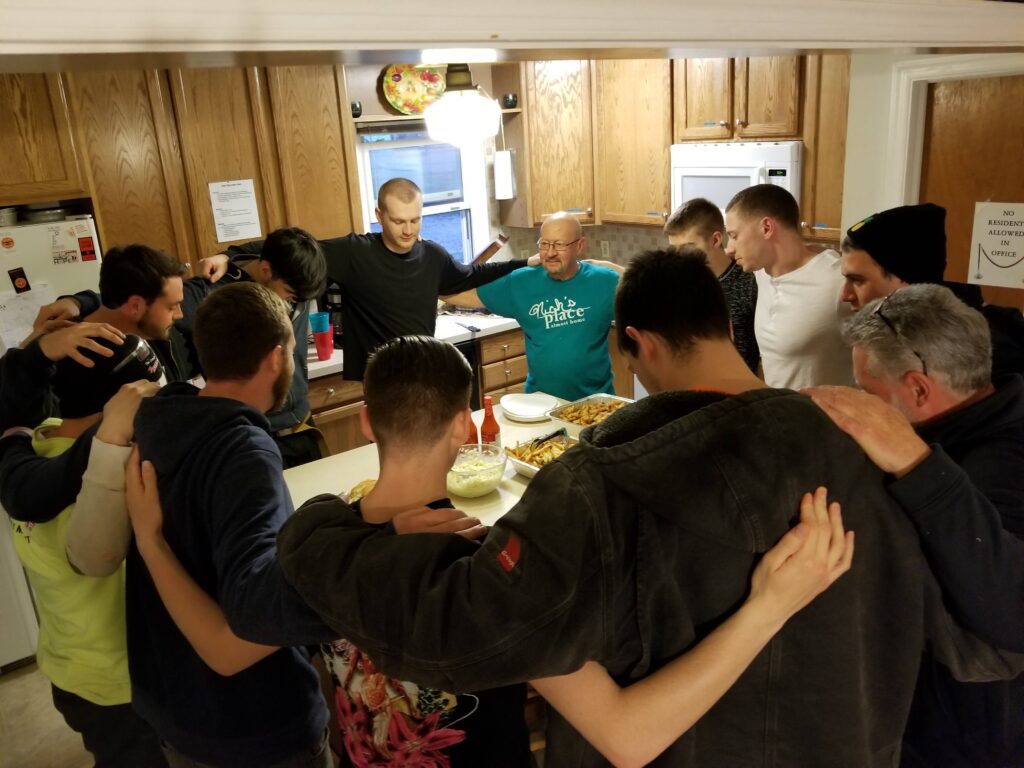 Twelve men stand in a circle around a kitchen island with their arms on each other's shoulders with their heads bowed, as if in prayer