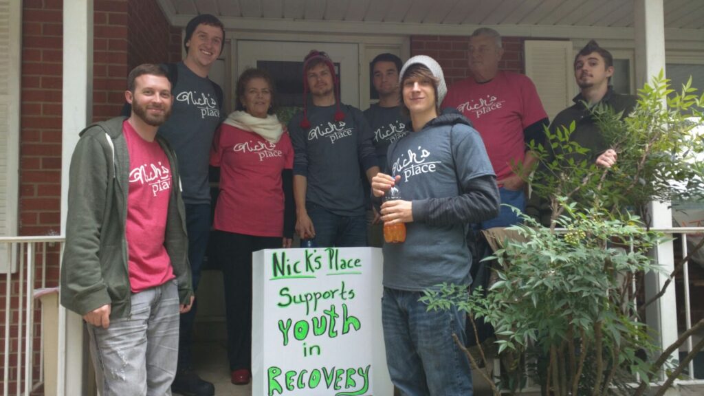 A group of people all dressed in red or gray matching "Nick's Place" t-shirts stand on the outside porch of a home around a hand-drawn sign that reads "Nick's Place Supports Youth in Recovery"