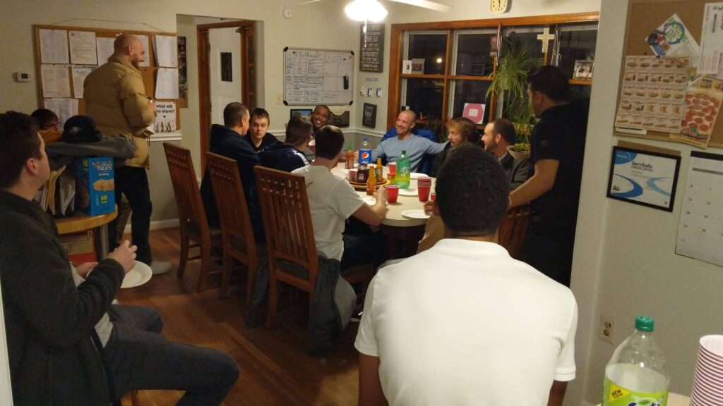 Ten young men, many smiling and laughing, are seated around a large dining table in a home while others sit on chairs or move around the room off to the side