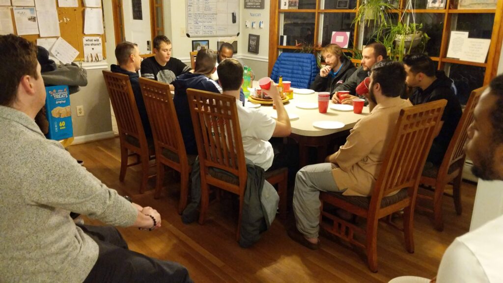 Ten young men gather around a large dining table in a home while others sit on chairs off to the side