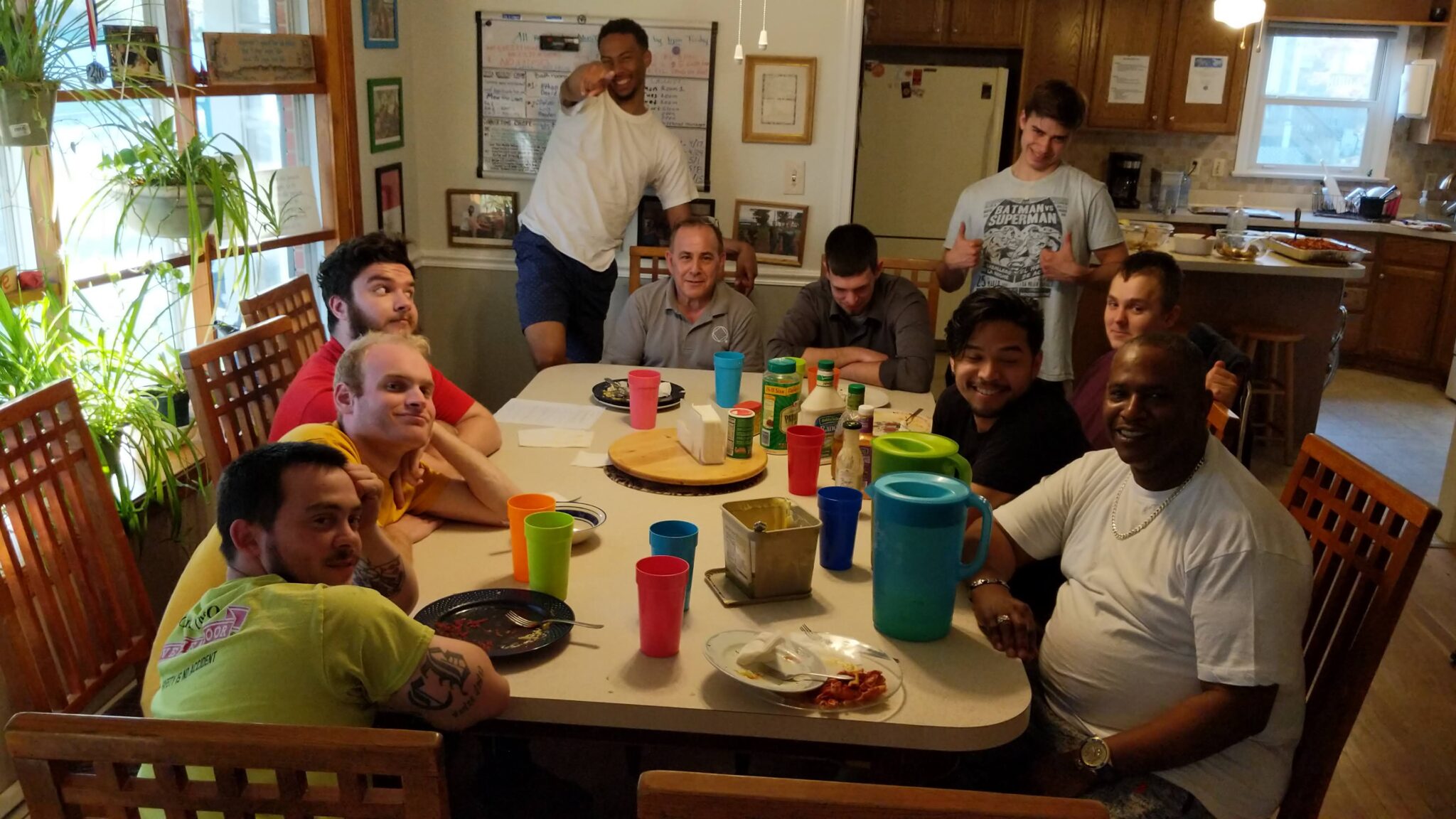 A group of ten men gather around a dining table of a home after sharing a meal, most are seated and smiling and others are standing and posing for the photo